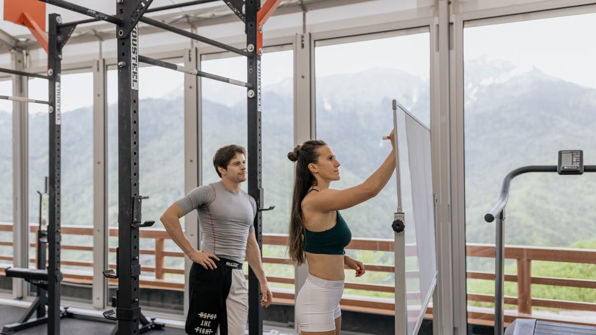 A gym coach instructs on whiteboard beside fitness equipment with mountain view.